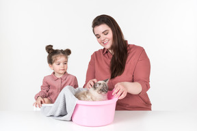 Мать и ее маленькая дочь в красных и розовых одеждах сидят за обеденным столом со своей семейной кошкой