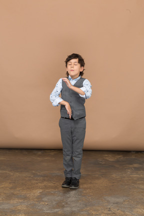 Vista frontal de un chico lindo en traje de pie con los ojos cerrados y gesticulando