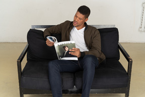 Schöner junger mann, der auf einem sofa sitzt und eine zeitschrift hält holding