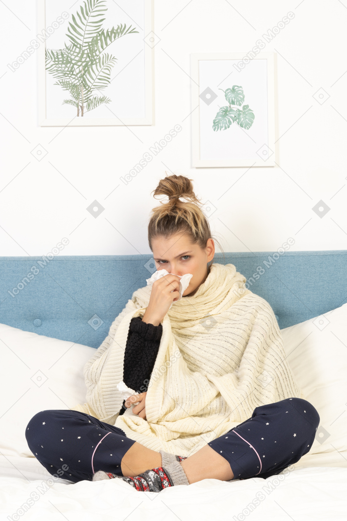 Front view of a young lady wrapped in white blanket sitting in bed and blowing nose