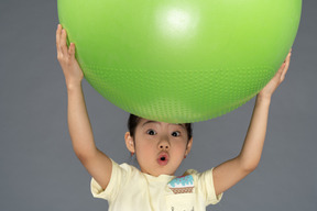 Menina surpresa segurando uma fitball verde acima da cabeça