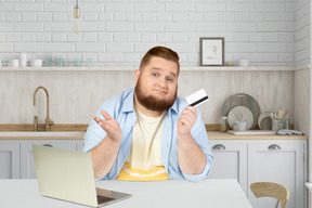A bearded man holding a credit card in front of a laptop in the kitchen