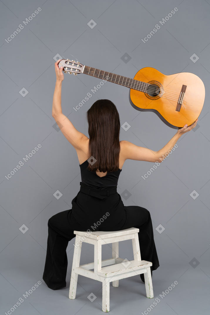 Young woman holding a guitar above her head while sitting backwards to camera