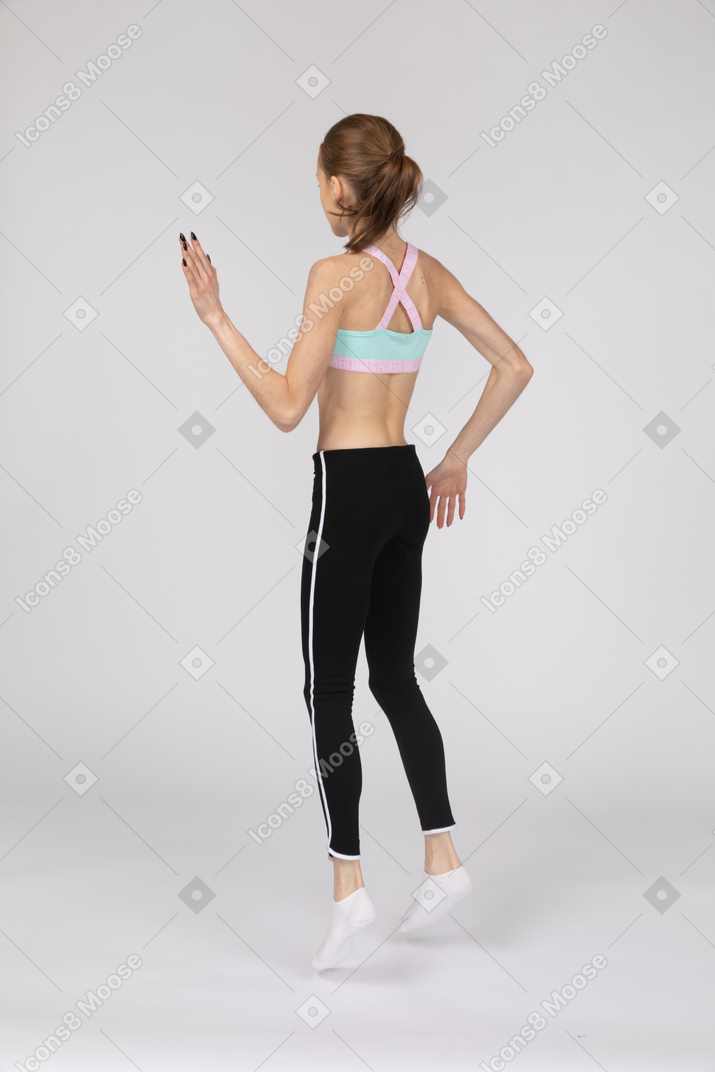 Vista traseira de três quartos de uma adolescente em roupas esportivas levantando as mãos e pulando