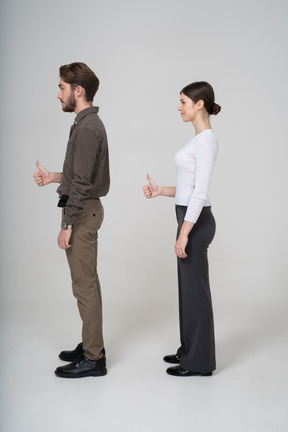 Vista lateral de una joven pareja alegre en ropa de oficina mostrando el pulgar hacia arriba
