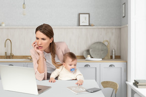 A woman sitting at a table looking at laptop with a baby on her lap