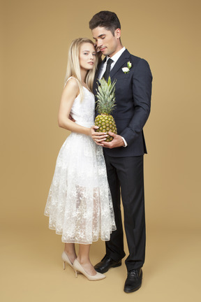 Noiva e noivo em pé ombro a ombro e segurando um abacaxi