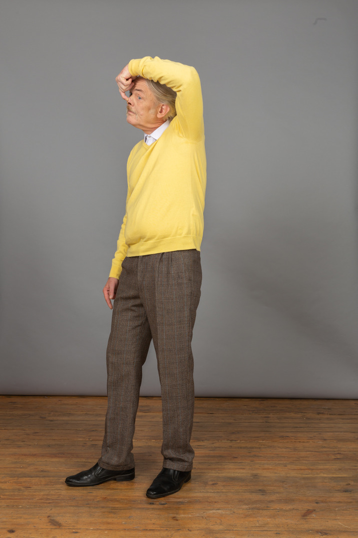Three-quarter view of an old funny man in yellow pullover touching nose and looking aside