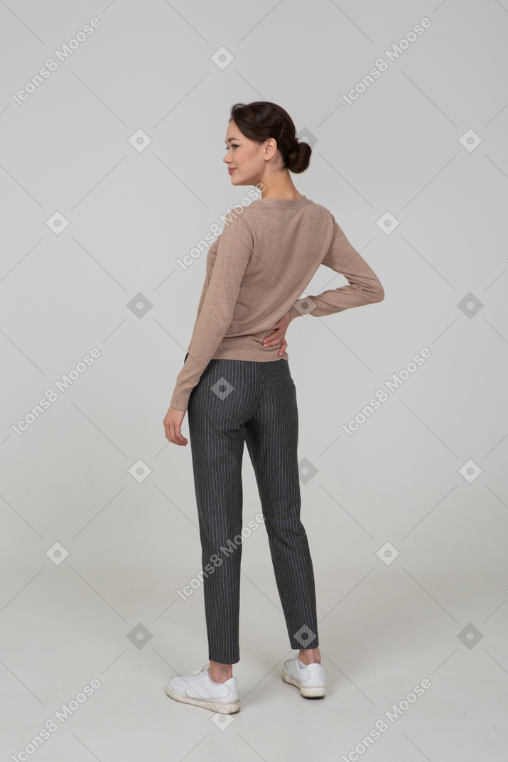 Three-quarter back view of a pleased female in pullover and pants putting hand on hip