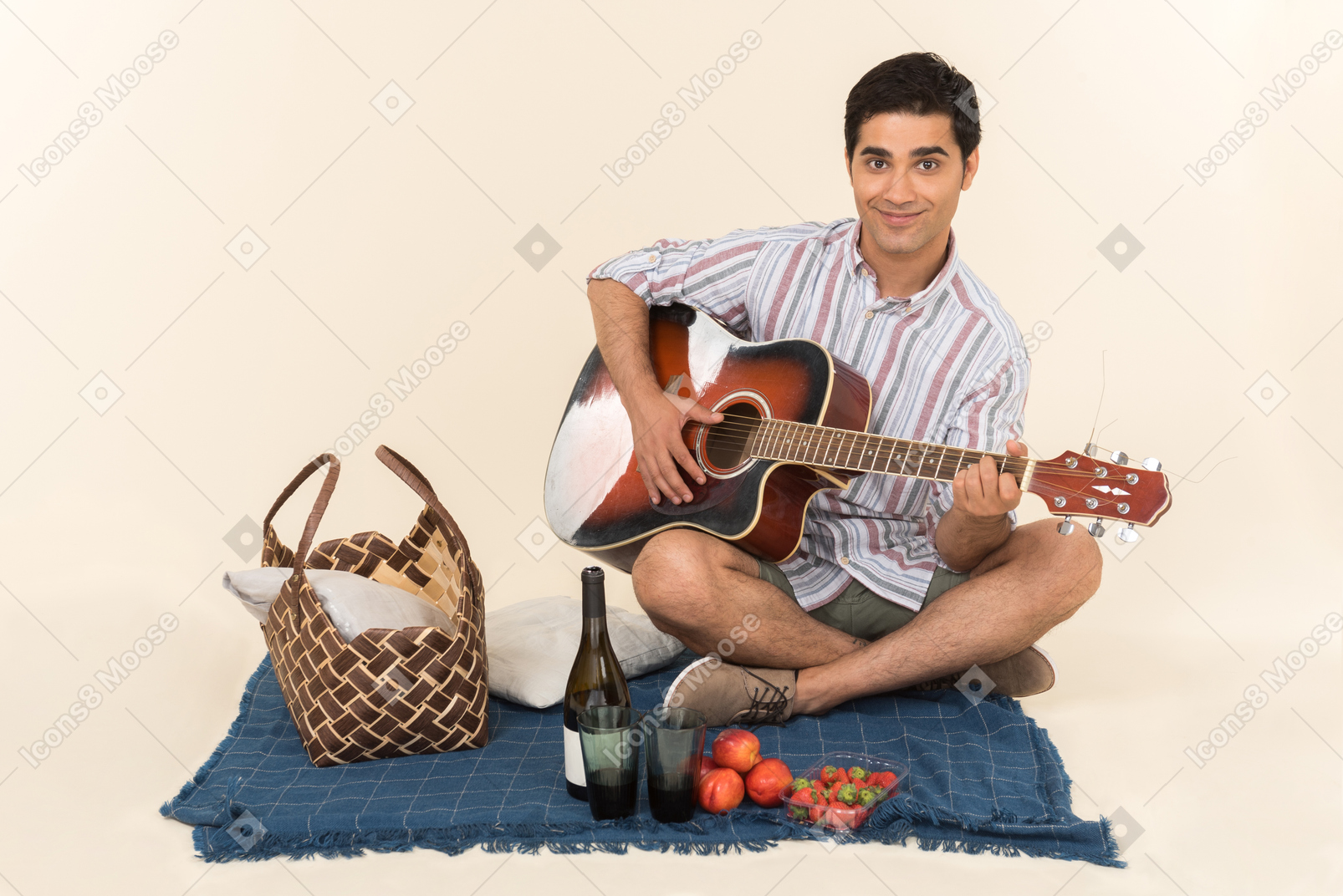 毛布の上のピクニックバスケットの近くに座って、ギターを弾く若い白人男