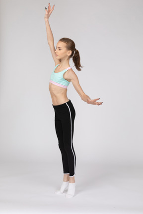 Вид в три четверти девушки-подростка в спортивной одежде, поднимающей руку и стоящей на цыпочках