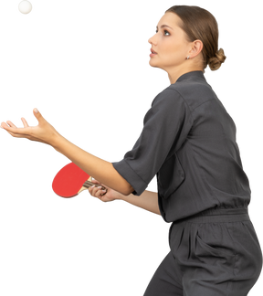 Side view of young woman in a jumpsuit serving tennis ball