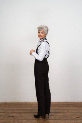 Vue latérale d'une vieille femme heureuse vêtue de vêtements de bureau mettant les mains ensemble en regardant la caméra