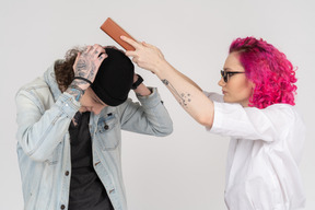 Niña de cabello rosado con gafas golpeando a su compañera de clase con un libro