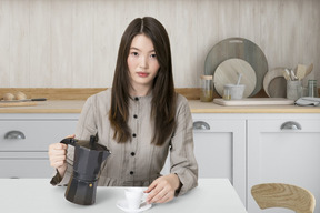 A woman sitting at a table holding a moka pot and a cup