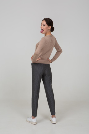 Vista posteriore di tre quarti di una giovane donna in pullover e pantaloni che mette le mani sui fianchi e mostra la lingua