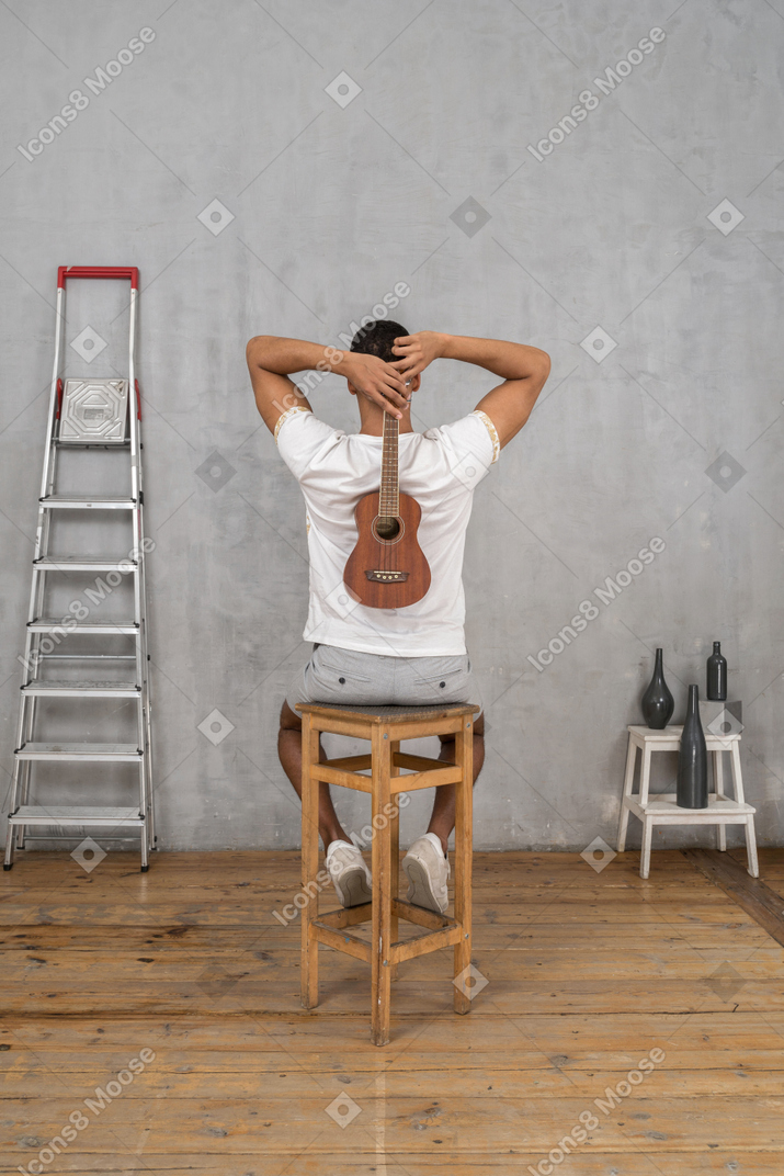 Back view of a man sitting on a stool holding an ukulele behind his back
