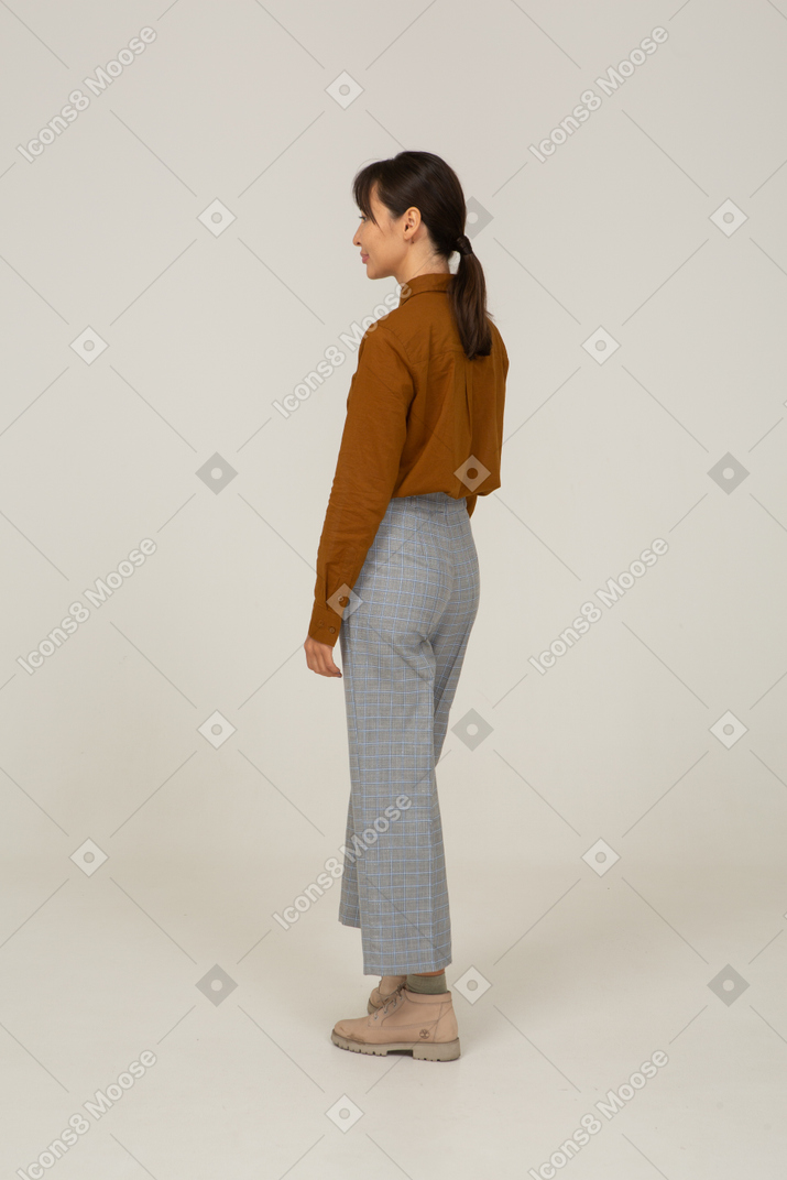 Vista traseira de três quartos de uma jovem mulher asiática fazendo beicinho, de calça e blusa, parada