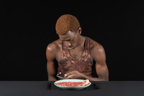 Vista frontal de um jovem afro espiando em seu telefone antes de comer carne