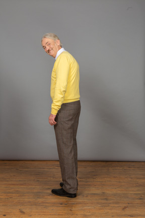 Three-quarter back view of an old man wearing yellow pullover and smiling while looking at camera