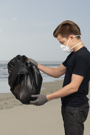 A man wearing a face mask and gloves holding a bag of garbage on a beach