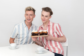 Pareja gay sentada en la mesa y sosteniendo una bandeja con cupcakes