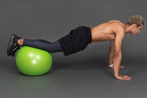 Vista lateral de un hombre afro sin camisa haciendo flexiones en una pelota de gimnasia