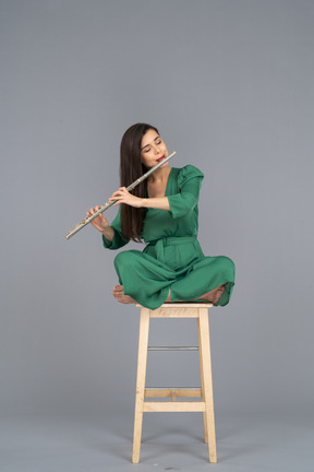 Toute la longueur d'une jeune femme jouant de la clarinette assise, les jambes croisées sur une chaise en bois