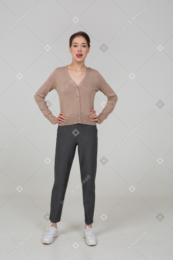 Front view of a young lady in pullover and pants putting hands on hips and showing tongue