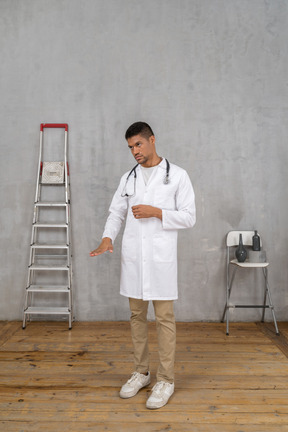 Three-quarter view of a young doctor standing in a room with ladder and chair showing a size of something