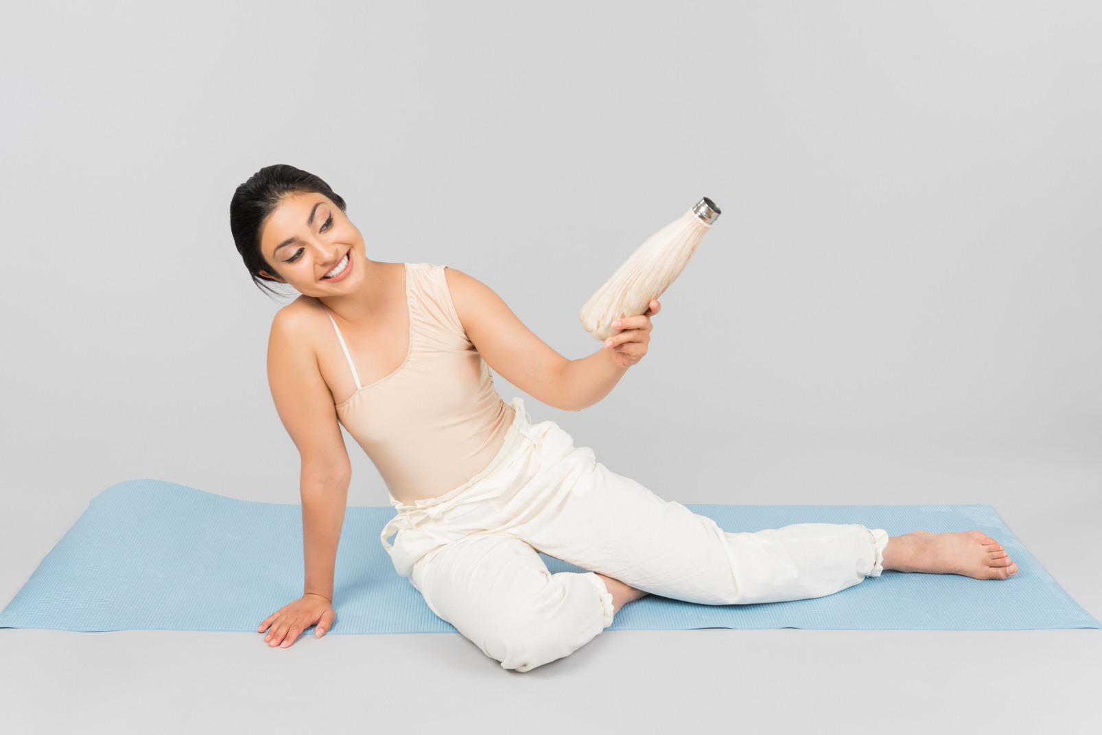 Young indian woman sitting on yoga mat and holding sport bottle