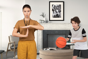 A female coach showing time out hand gesture next to a boy with basketball ball in a living room