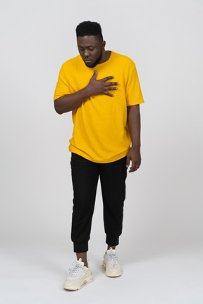 Front view of a surprised young dark-skinned man in yellow t-shirt touching chest