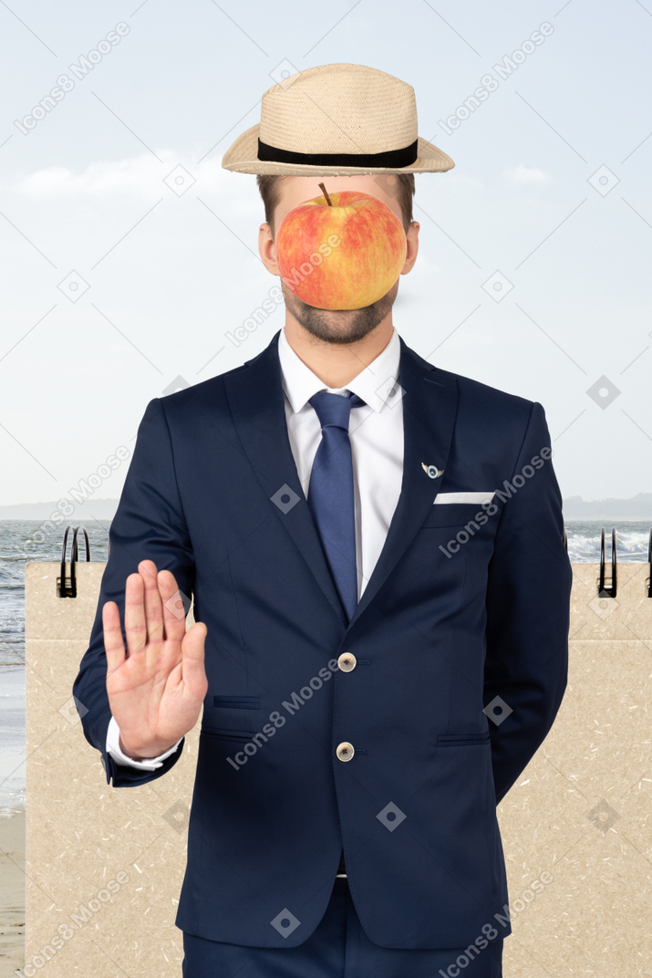 Um homem de chapéu com uma maçã no rosto em pé na frente de um notebook perto do mar com a palma da mão para a câmera