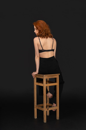 A back side of the beautiful woman dressed in black pants and bra, sitting on the wooden chair and looking to the left