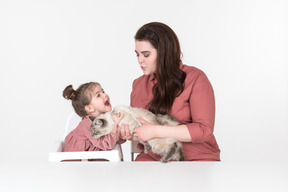 Madre y su pequeña hija, vestidas de rojo y rosa, sentadas a la mesa con su gato familiar