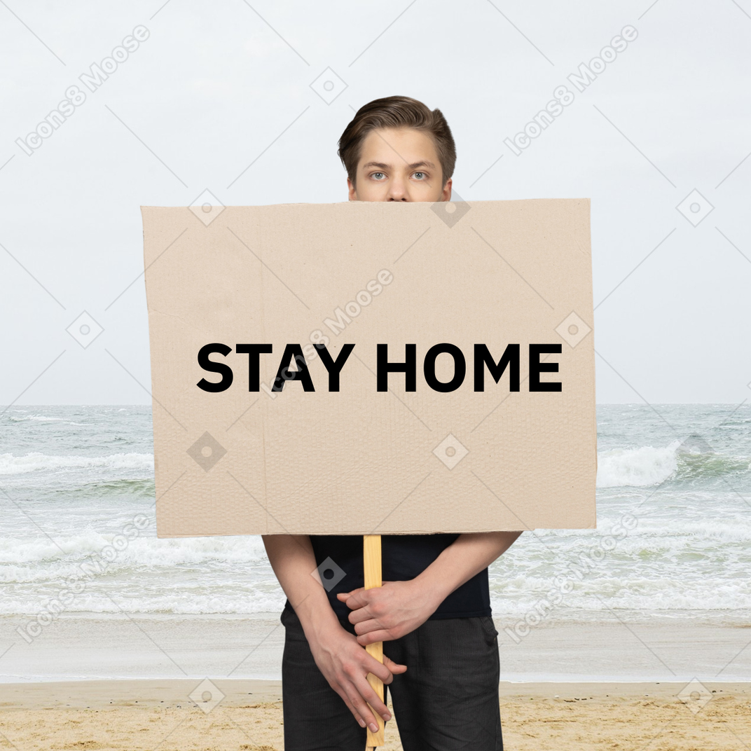 집에 머무르는 표지판을 들고 해변에 서있는 남자