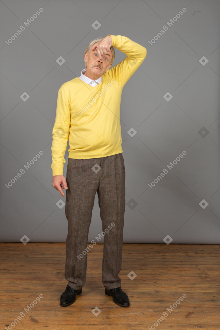 Front view of an old funny man in yellow pullover touching nose and looking at camera