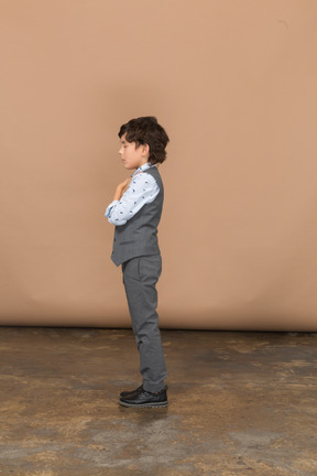 Vista lateral de un niño con traje gris posando con los brazos cruzados