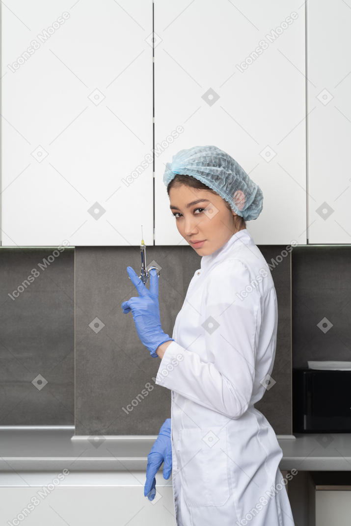 Side view of a female doctor in a medical hat holding a syringe and looking at camera