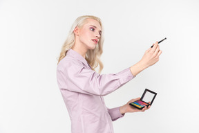 Jovem de cabelos loiros em uma camisa violeta pastel, fazendo a maquiagem de alguém