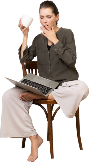 Vista frontale di una giovane donna scioccata che indossa abiti da casa seduta su una sedia con un laptop e beve caffè