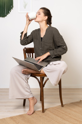 Vista frontal de una mujer joven vistiendo ropa de casa sentado en una silla con una computadora portátil y tomando café
