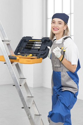 A woman in overalls holding a tool box on a ladder