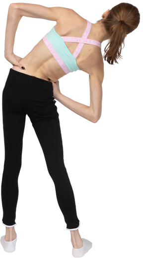 Vista posteriore di una ragazza adolescente in abbigliamento sportivo che mette le mani sui fianchi mentre si appoggia a destra