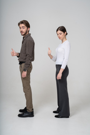 Vista lateral de um jovem casal alegre com roupas de escritório aparecendo o polegar