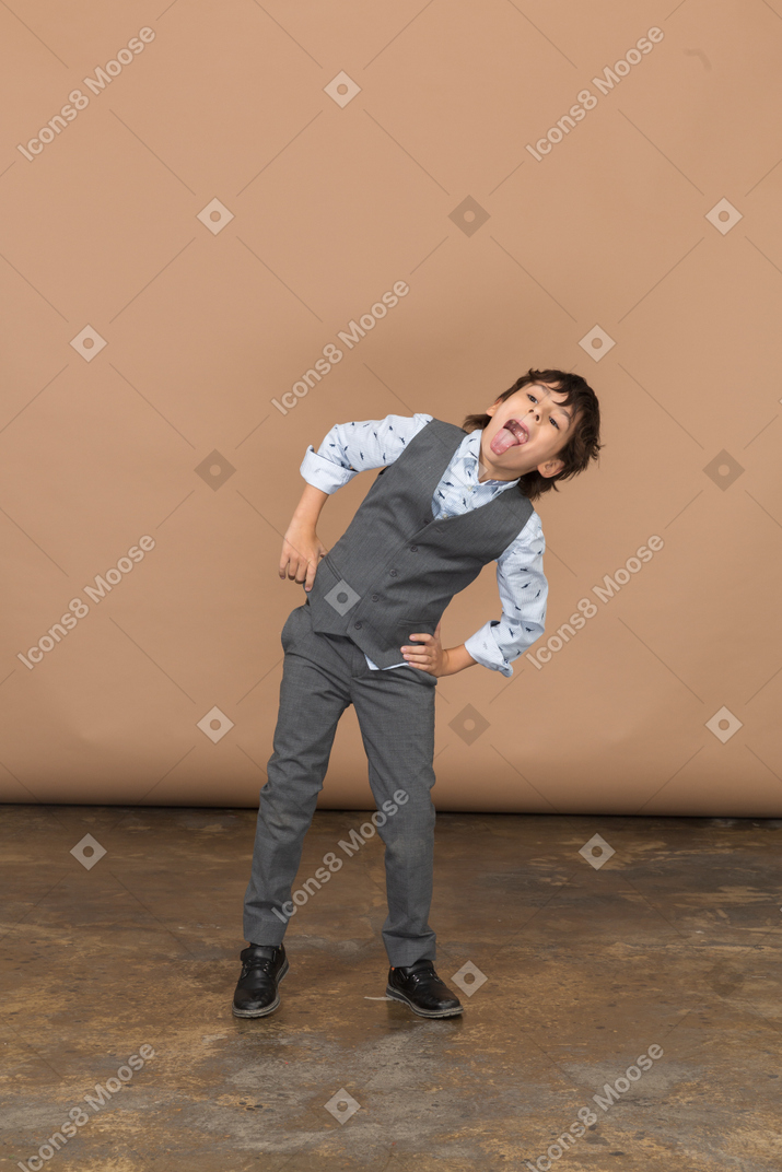 Vista frontal de um menino de terno posando com as mãos nos quadris e mostrando a língua