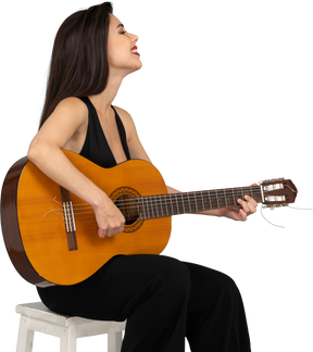 Вид сбоку сидящей улыбающейся молодой леди в черном костюме, играющей на гитаре