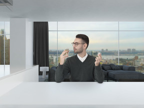 Handsome young man in a grey sweater and round glasses, smelling a bottle of a new perfume in a spacious modern-looking room