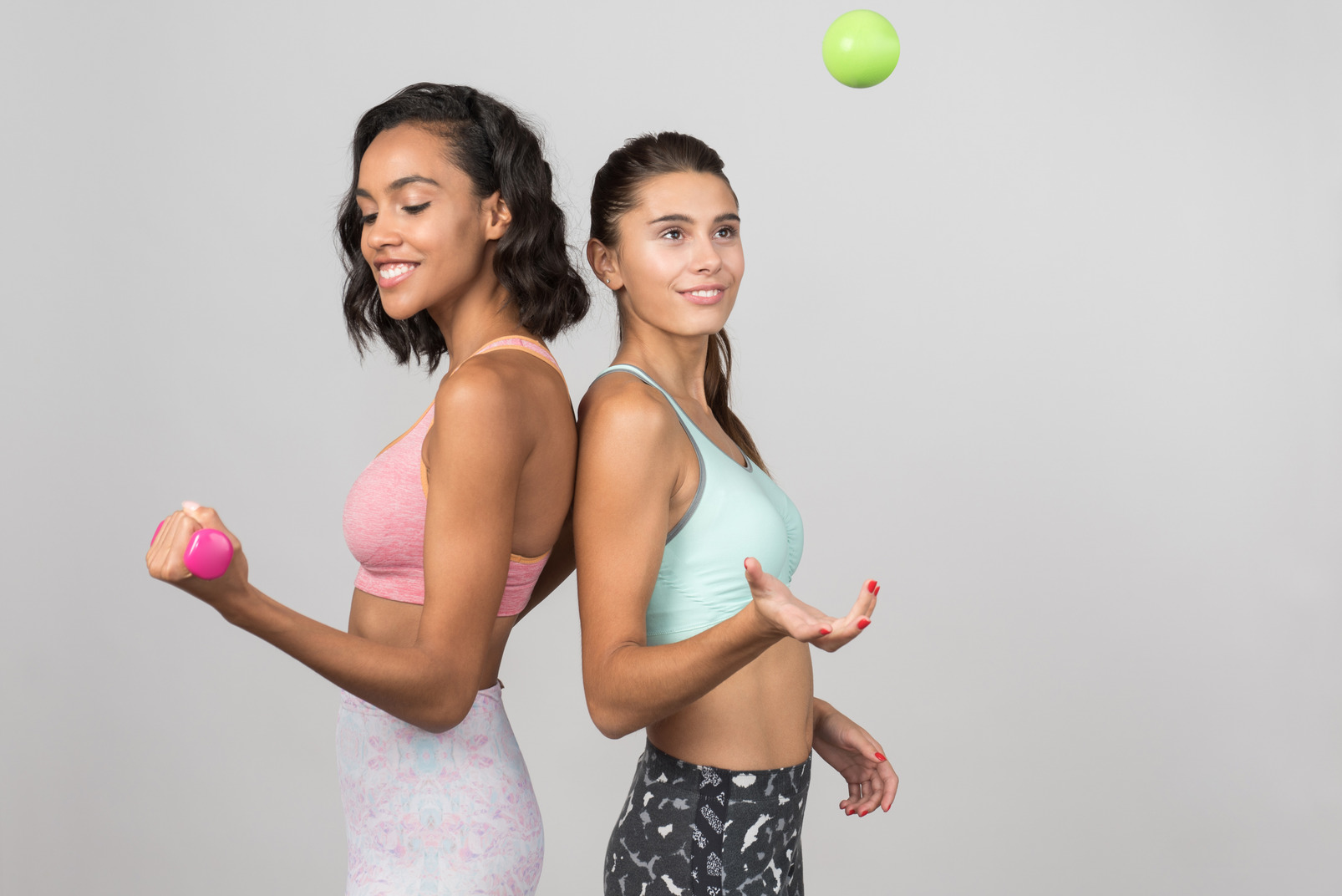 Two young girls standing shoulder to shoulder and one is holding hand weights and another one throwing tennis ball